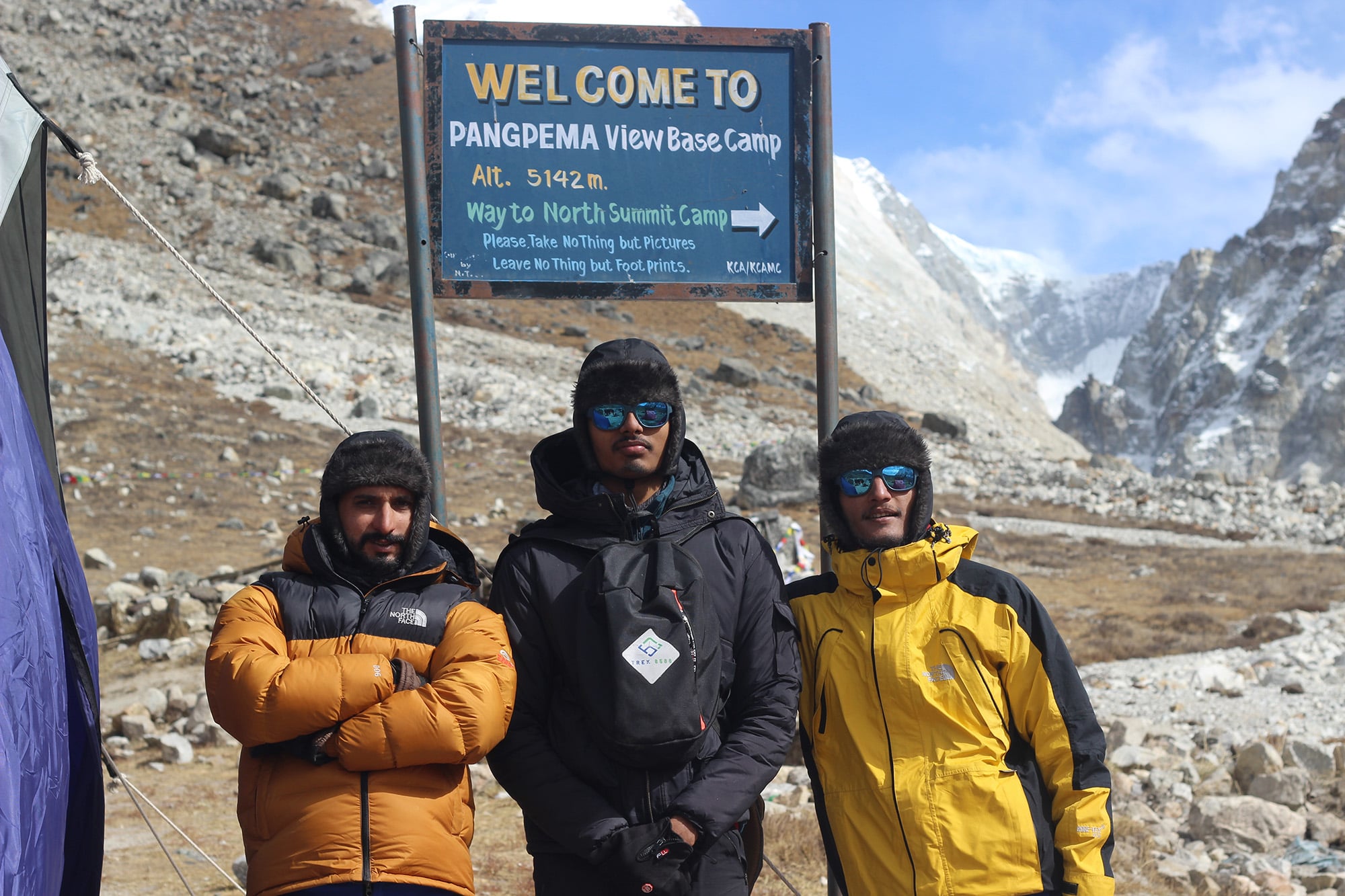 Three guides posing in front of a banner featuring Pangpema Base Camp, Kanchenjunga Region, symbolizing expert guidance for trekking adventures in Eastern Nepal.