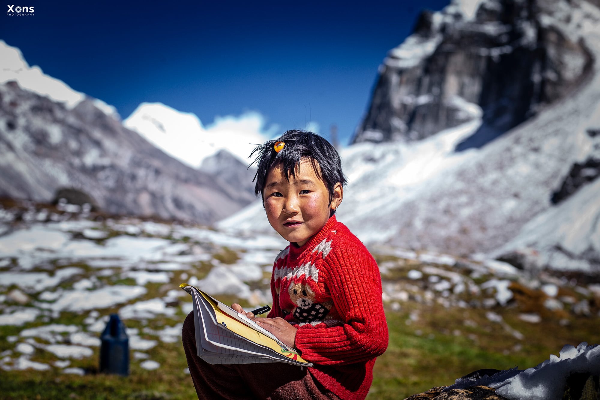 A small boy reading in front of the majestic mountains in Khambachen, Kanchenjunga Region, capturing a peaceful moment amidst the stunning natural beauty of Eastern Nepal.